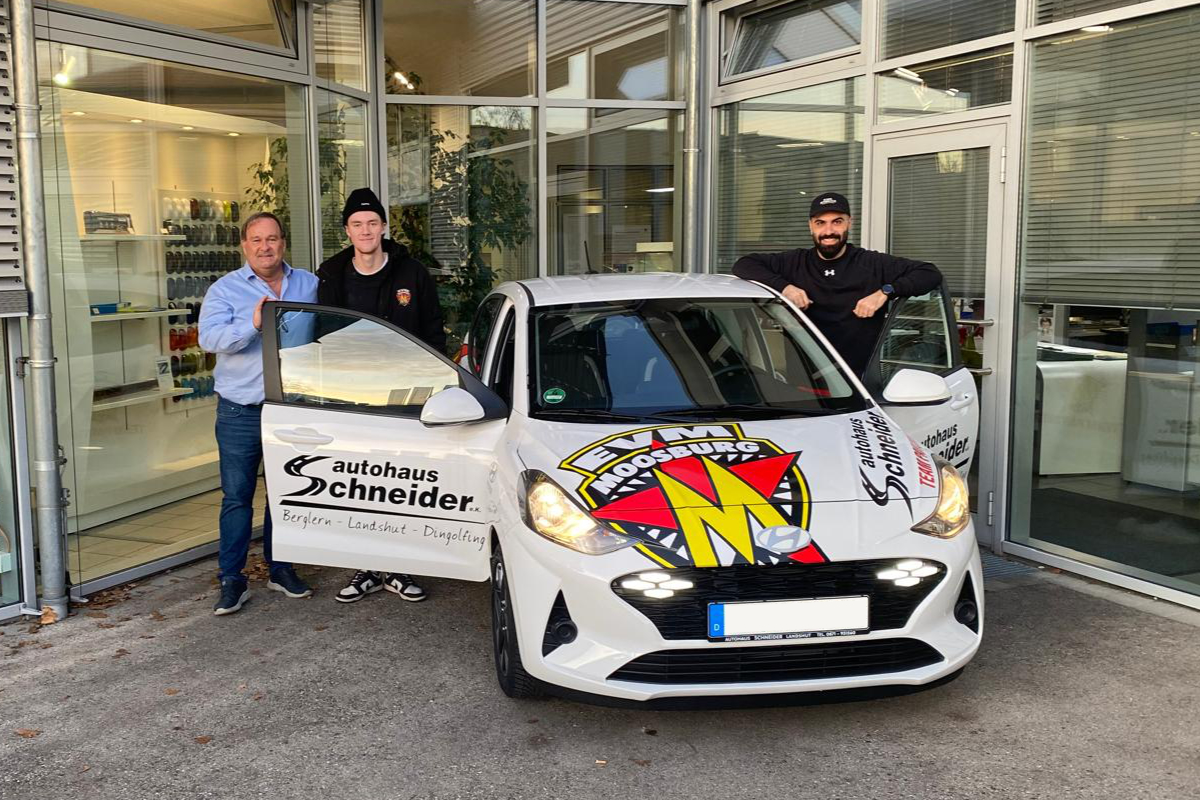 Drei Männer stehen vor einem Autohaus neben einem weißen Auto mit „EV Moosburg“-Beklebung.